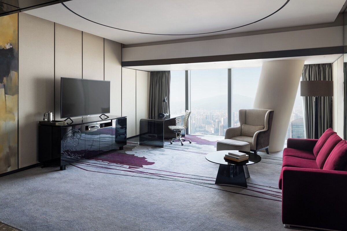 Et moderne hotelværelse med et stort vindue der giver udsigt over byen, et fladskærms-tv, et skrivebord med stol, en rød sofa og et sofabord.