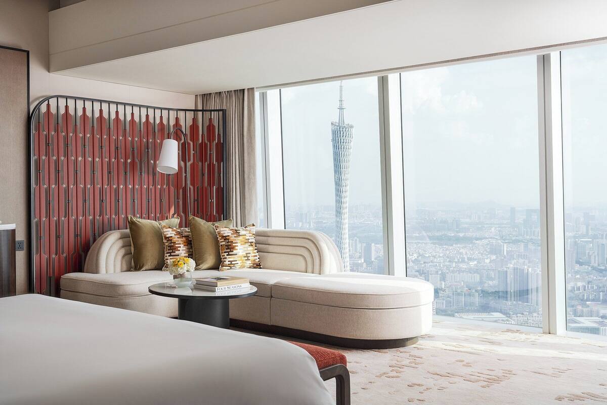 A modern hotel room with a large window offering a city view, a sofa with pillows, a bedside table, and a lamp.