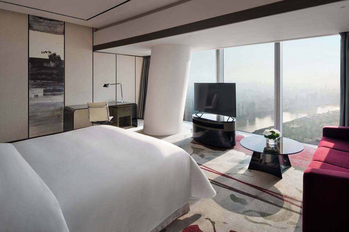 A modern hotel room with a large bed, a red sofa, a TV, and a city view.