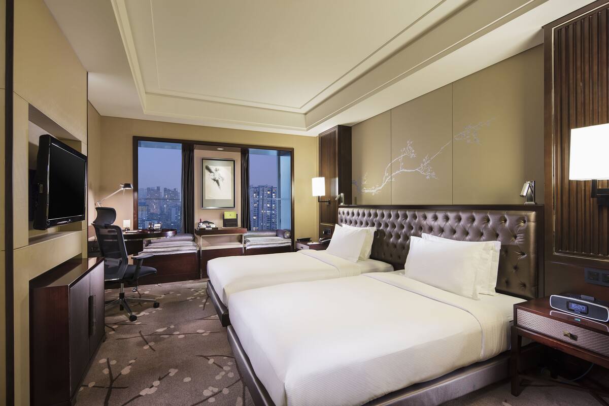Ein Hotelzimmer mit zwei Betten, einem Schreibtisch mit Computer, einem Fernseher und Blick auf die Stadt.