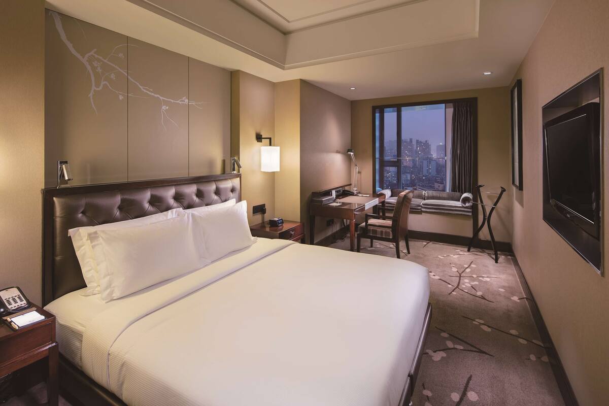 Ein Hotelzimmer mit einem großen Bett, einem Schreibtisch, einem Sessel, einem Fernseher und einem Fenster mit Blick auf die Stadt.