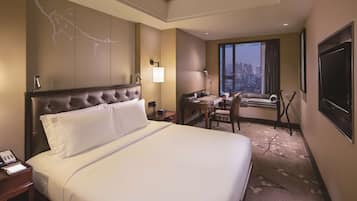 A hotel room with a large bed, a desk, a chair, a TV, and a window with a city view.