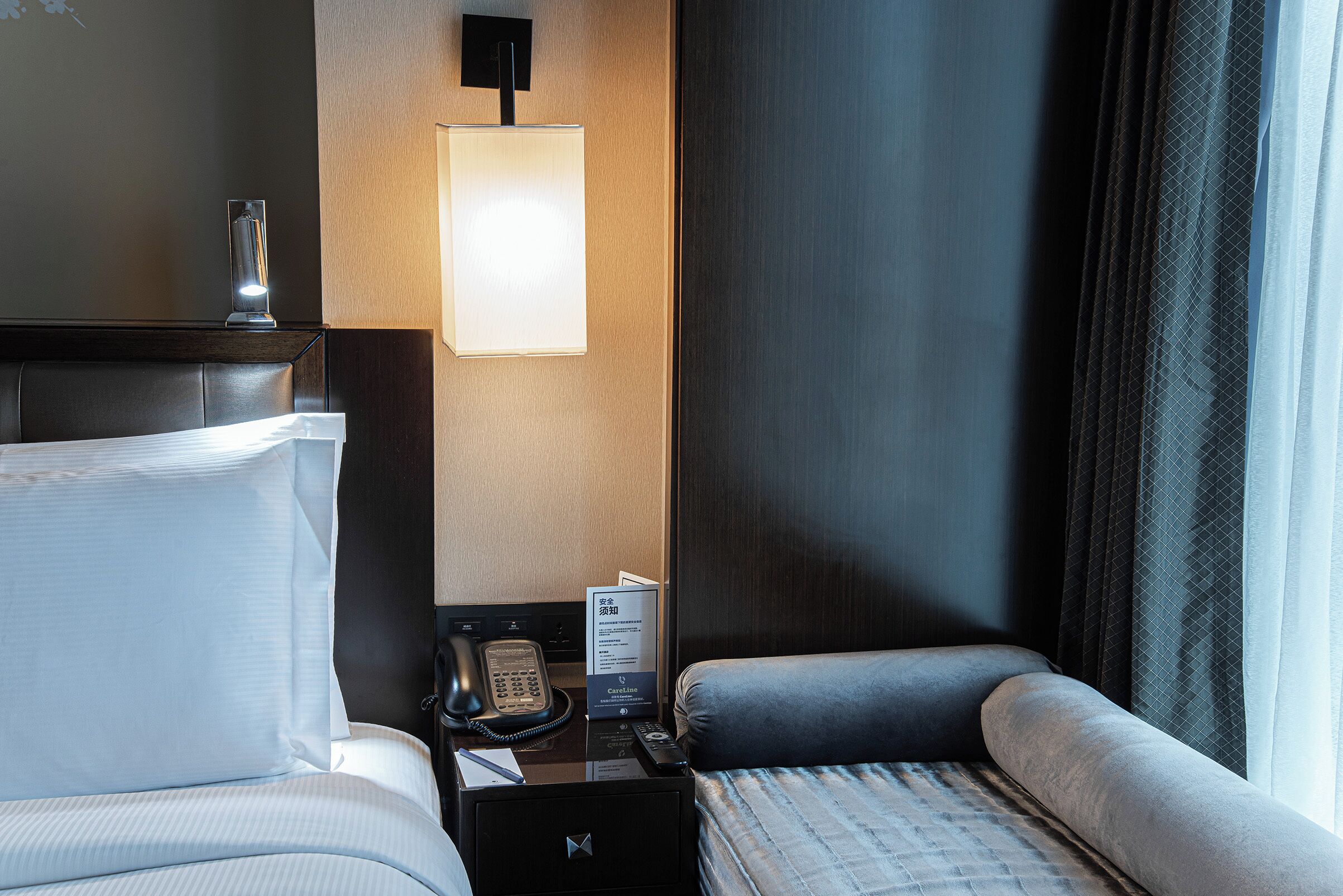 Habitación de hotel con cama, mesita de noche, lámpara y vistas al exterior a través de una ventana.