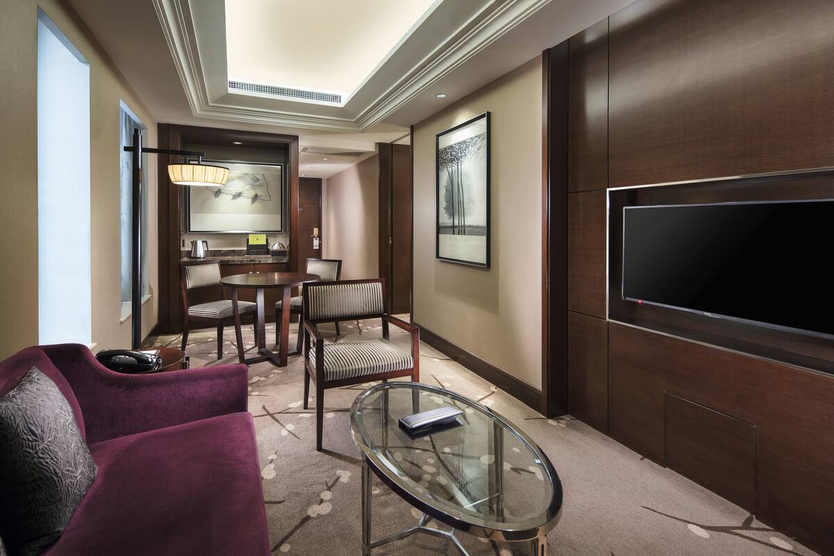 Ein modernes Hotelzimmer mit einem großen Flachbildfernseher, einem Glastisch, einem violetten Sessel und einem Essbereich mit Tisch und Stühlen.