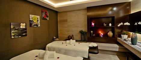 A spa room with two massage tables, a wall-mounted shelf with decorative items, and a ceiling light fixture.