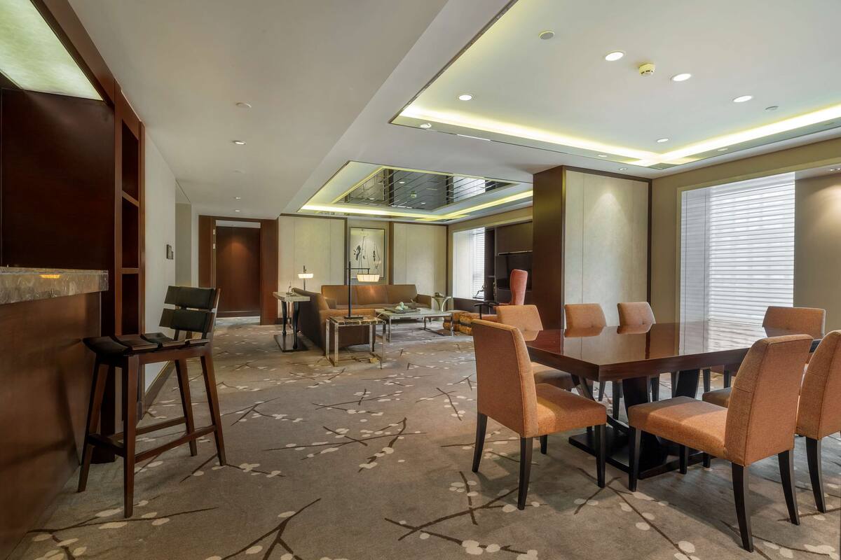 Ein modernes Hotelzimmer mit Essbereich, einer Couch, einem Couchtisch und einem großen Fenster mit Jalousien.