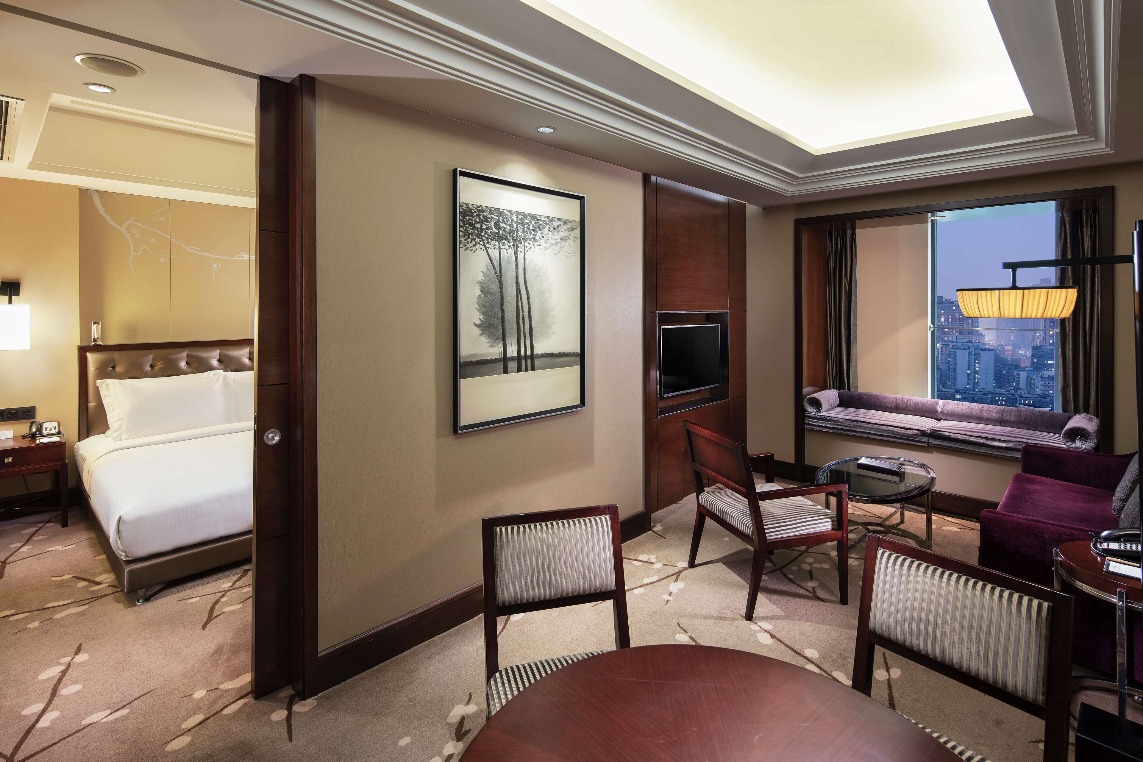A hotel room with a bed, a seating area with a sofa and chairs, a TV, and a large window with a city view.