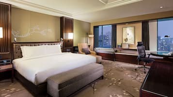 A hotel room with a large bed, a desk, a chair, and a view of the city at night.