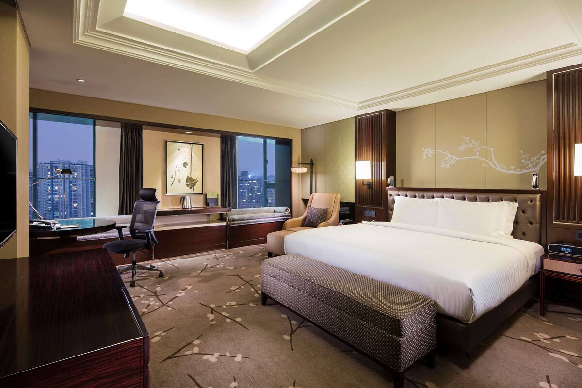 Ein Hotelzimmer mit einem großen Bett, einem Schreibtisch, einem Sessel, einem Fernseher und Blick auf die Stadt.
