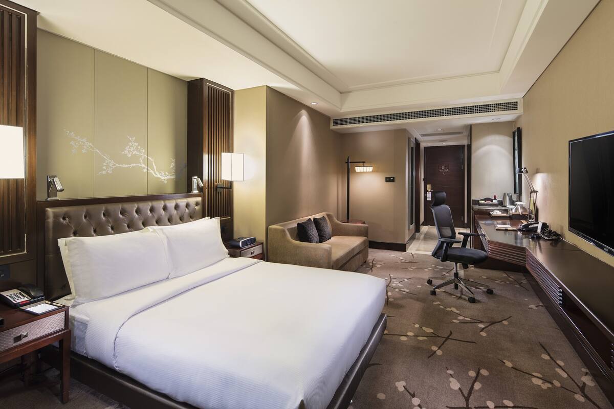 Ein Hotelzimmer mit einem großen Bett, einem Schreibtisch, einem Sessel, einer Couch, einem Fernseher und einem durchgegangenen Tür erkennbaren Badezimmer.