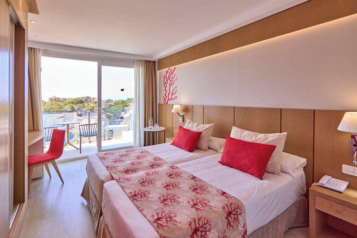 A hotel room with two beds, a red chair, a small table, and a view of the city.