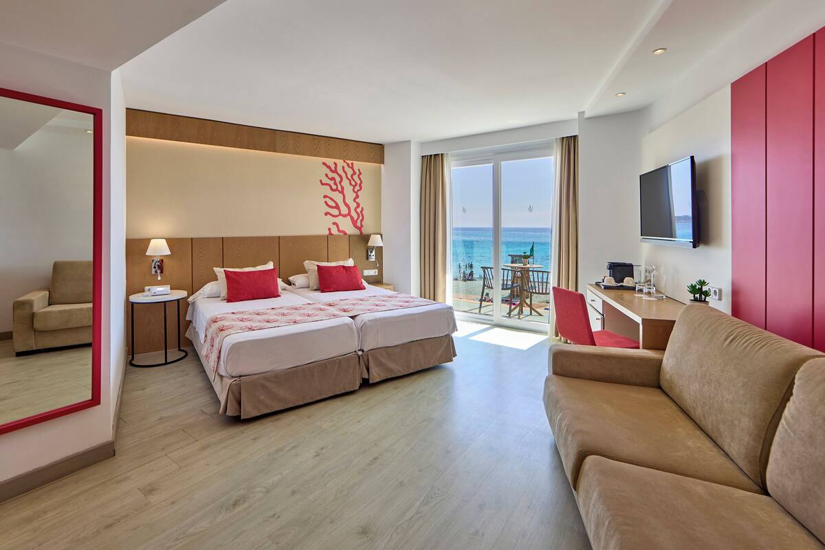 A modern hotel room with a large bed, a sofa, a TV, and a balcony with a view of the sea.