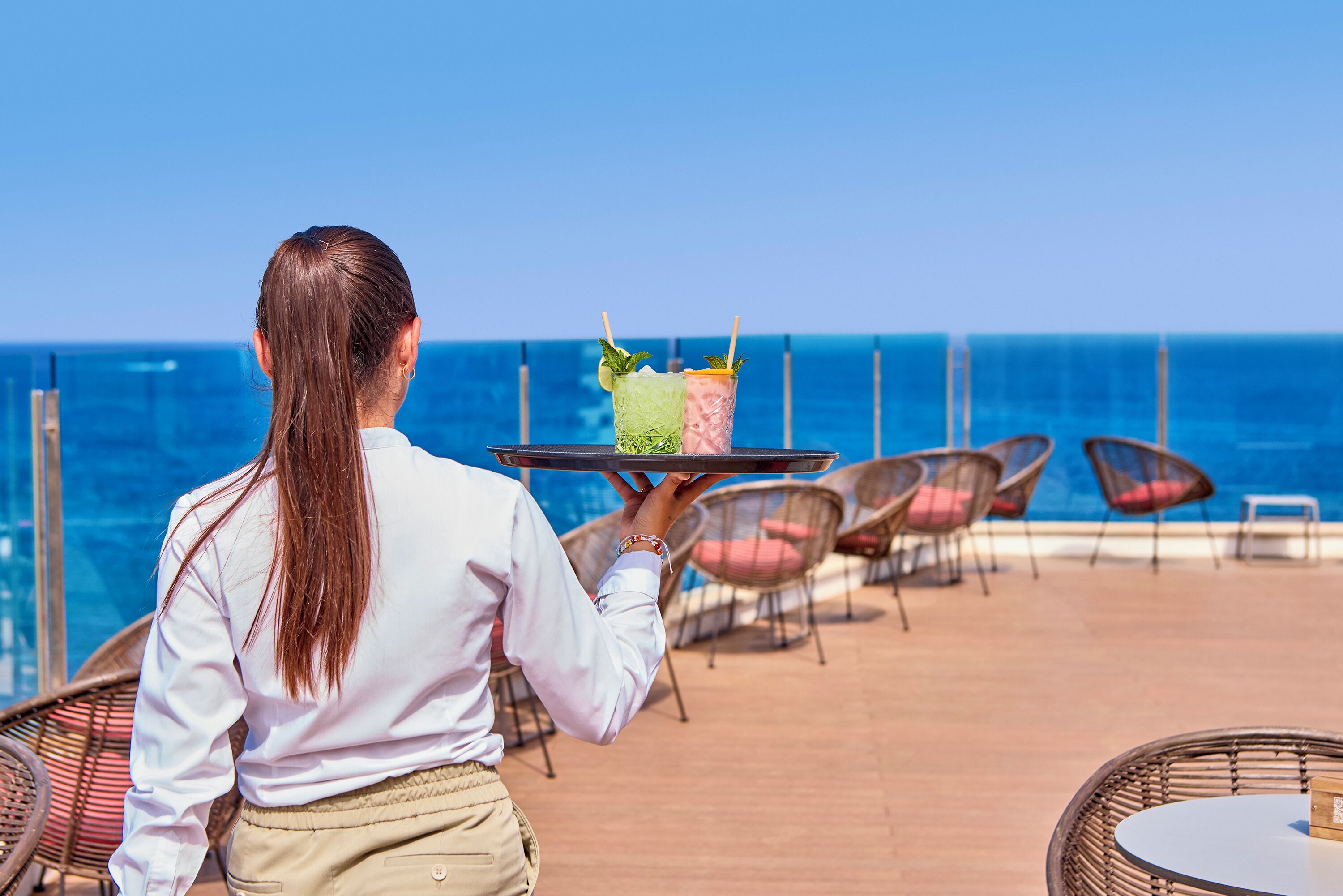 Una persona lleva una bandeja con dos bebidas en una terraza con sillas y vista al océano.