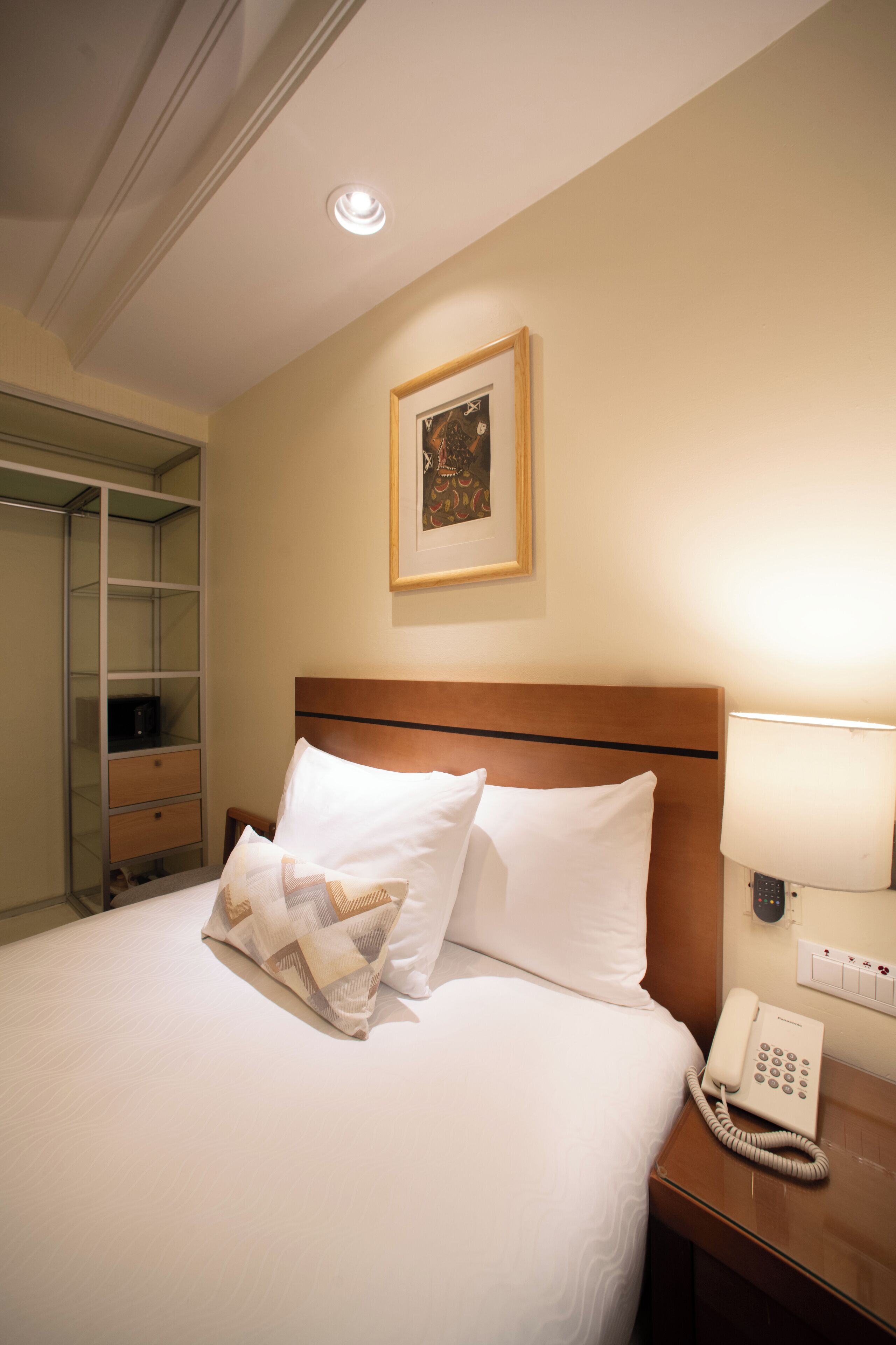 A hotel room with a bed, a bedside telephone, a lamp, a framed picture on the wall, and a closet with a mirror.