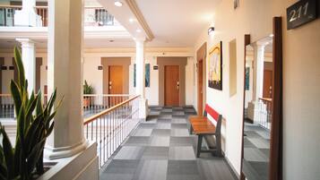 A hotel corridor with checkered flooring, a wooden bench, a potted plant, and a mirrored wall with a door number sign.