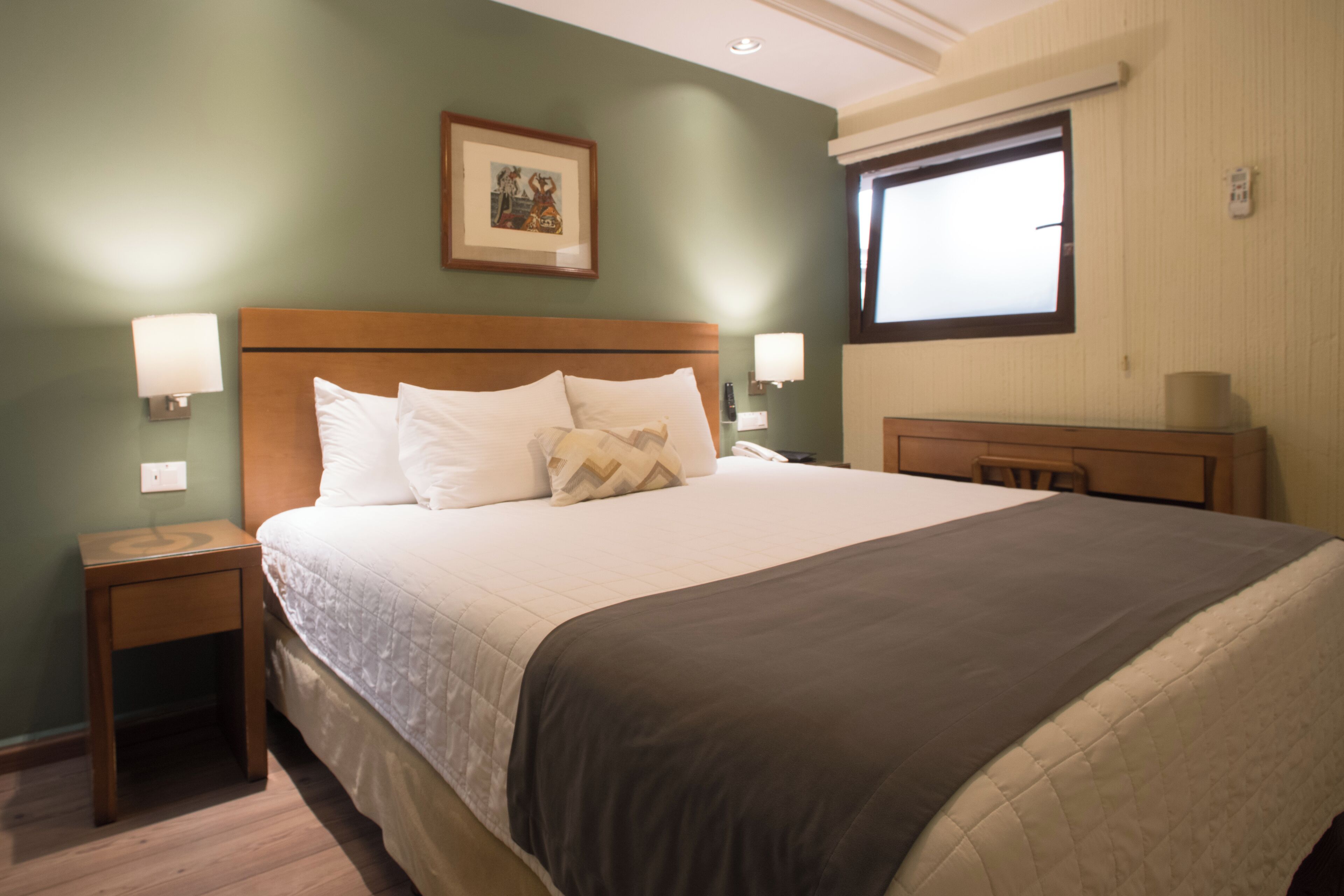 Una cama bien hecha con cabecera de madera, dos mesitas de noche con lámparas, un cuadro en la pared y una ventana con persianas.