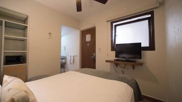 Una habitación de hotel con una cama, un televisor en un estante, un ventilador de techo y una puerta que da al baño.