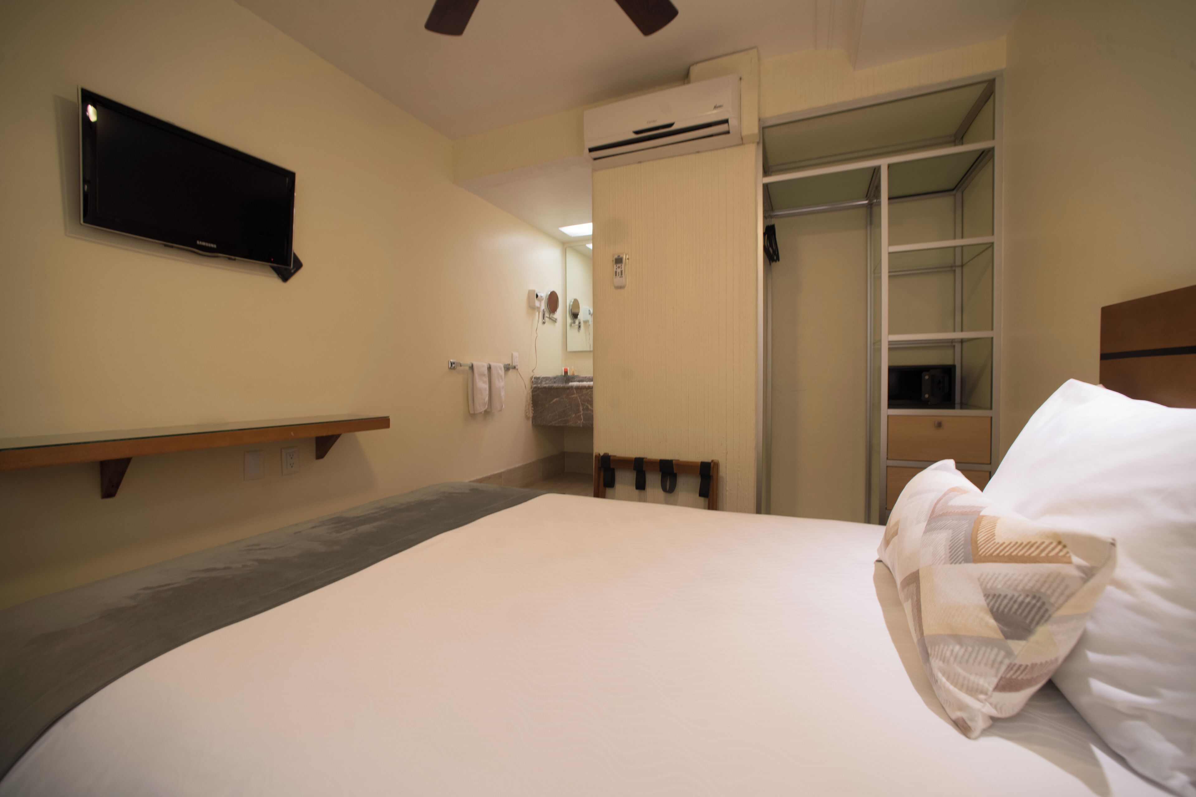 A hotel room with a bed, a flat-screen TV mounted on the wall, a wooden shelf, and a closet with a sliding door.