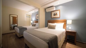 Una habitación de hotel con dos camas, piso de madera, ventilador de techo, aire acondicionado, una mesita de noche con una lámpara y un cuadro enmarcado en la pared.