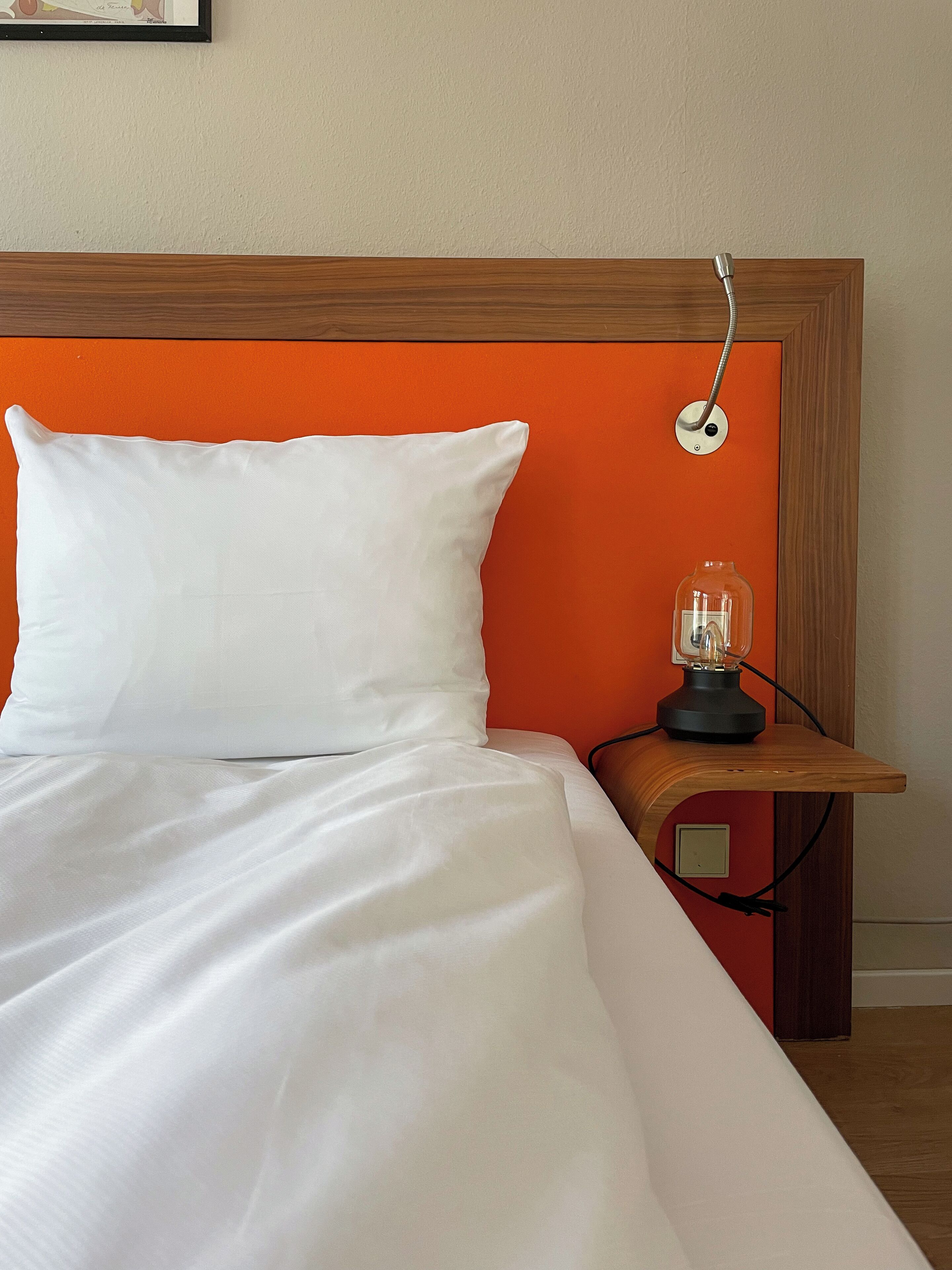 A neatly made bed with white linens, a wooden headboard with an orange panel, and a bedside table with a lamp.