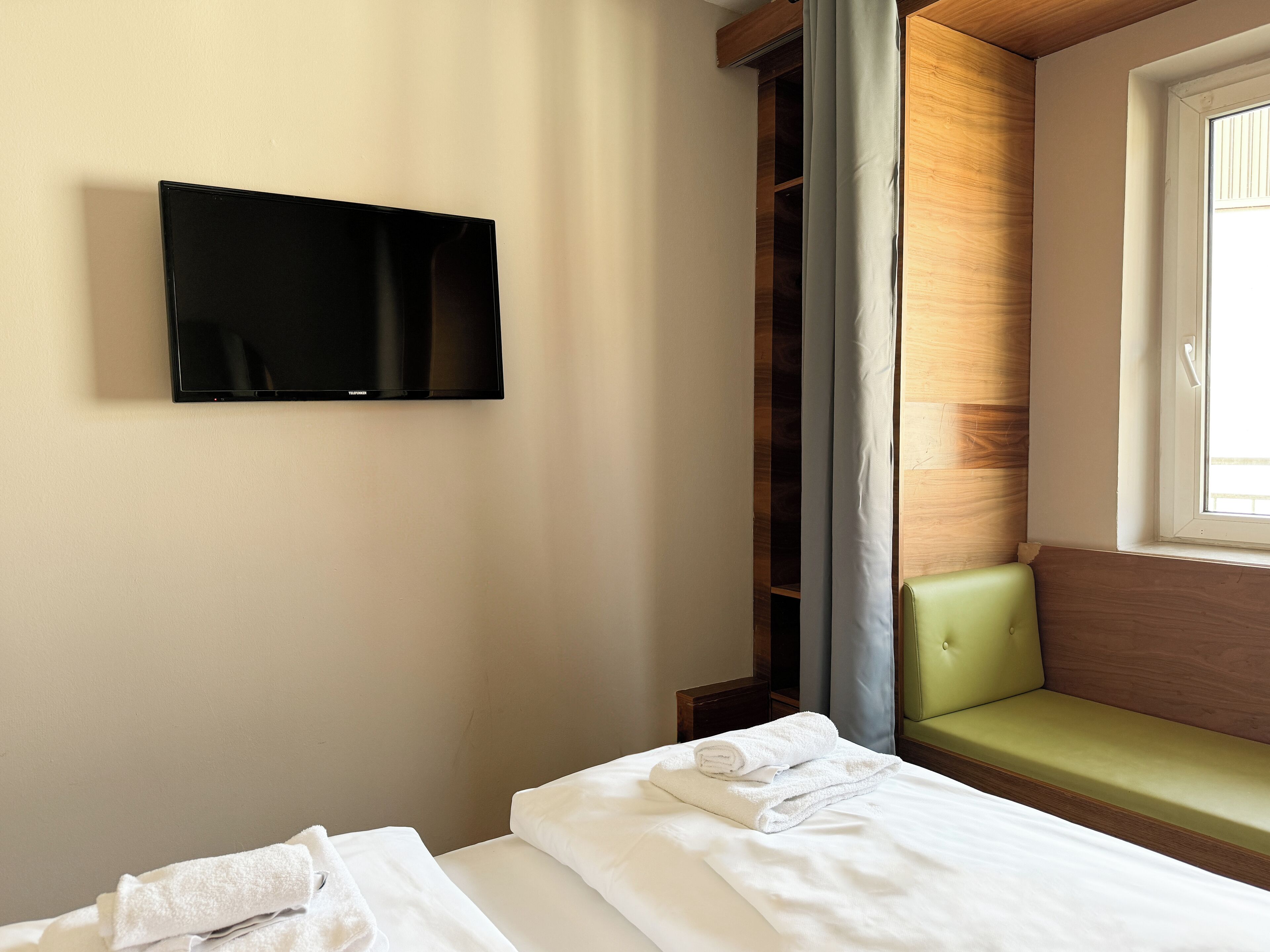 A hotel room with a bed, a television mounted on the wall, a window with a view, and a green cushioned bench.