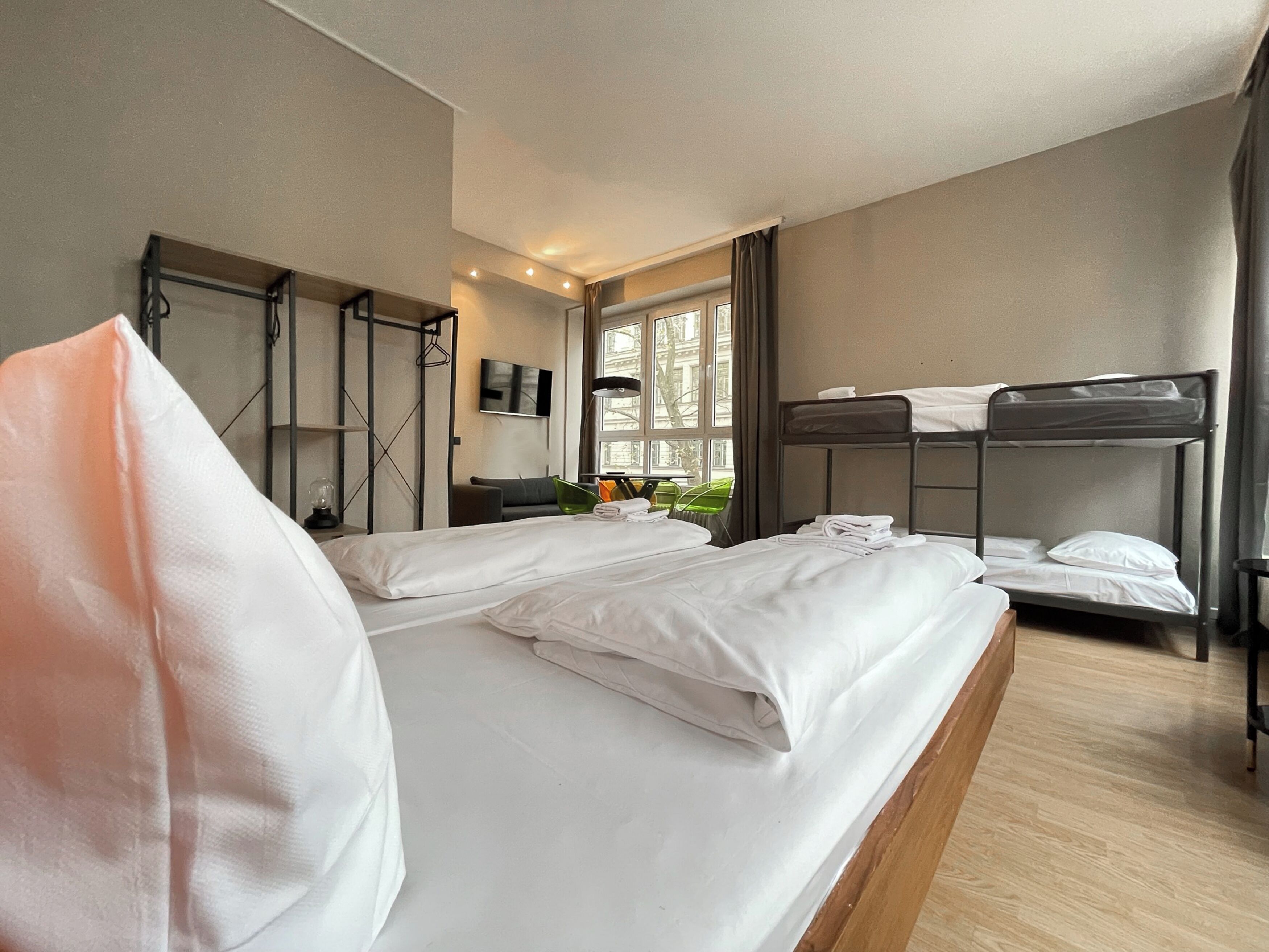 A hotel room with a large bed, a bunk bed, a TV, a window with curtains, and a small table with a lamp.