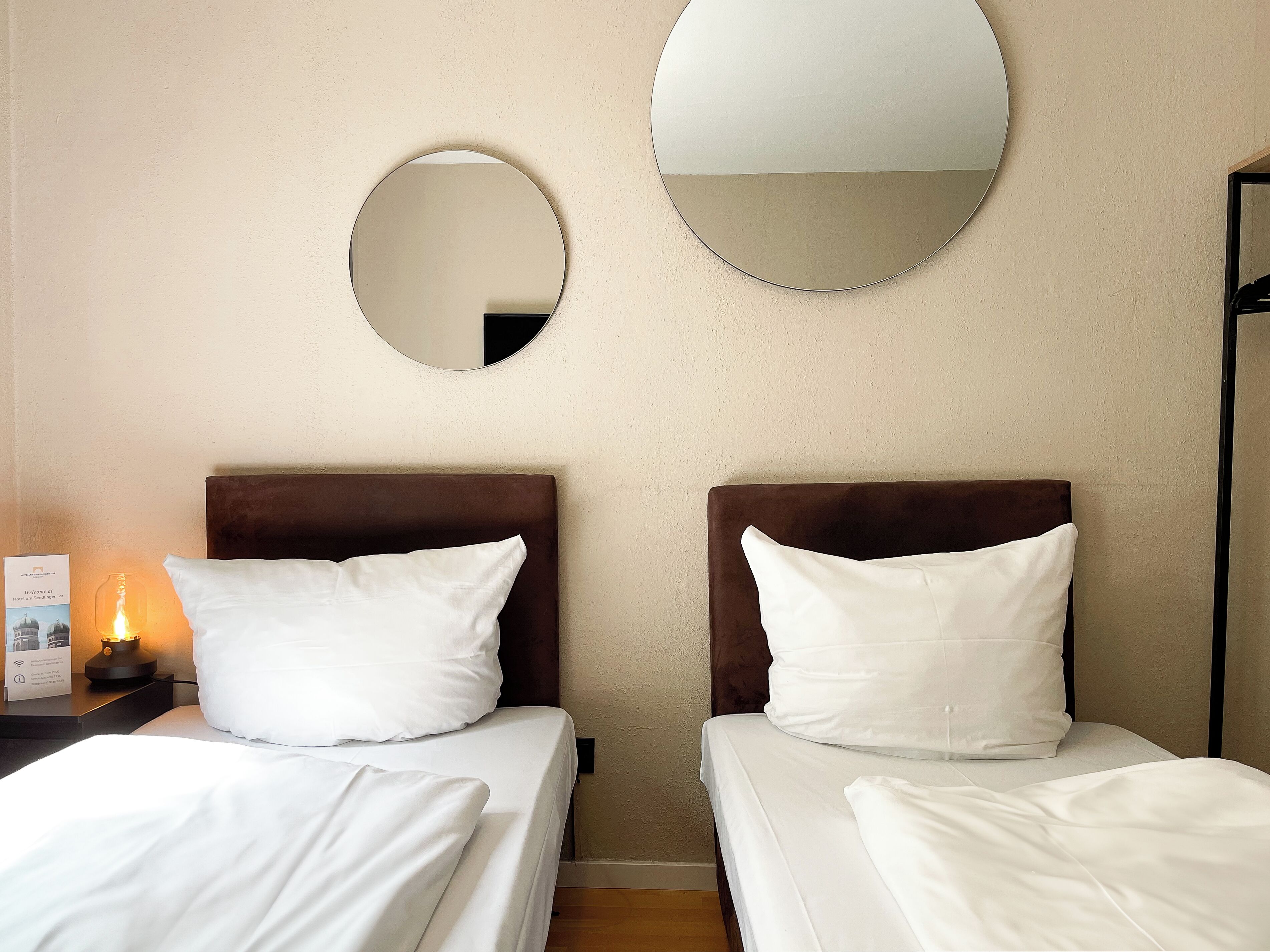 A hotel room with two beds, each with white bedding and brown headboards. There are two round mirrors on the wall, and a nightstand with a lamp and a sign.