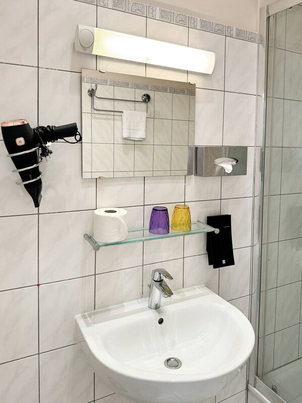 Un baño con un lavamanos blanco, un espejo, un perchero para toallas y un estante de vidrio con artículos de tocador.