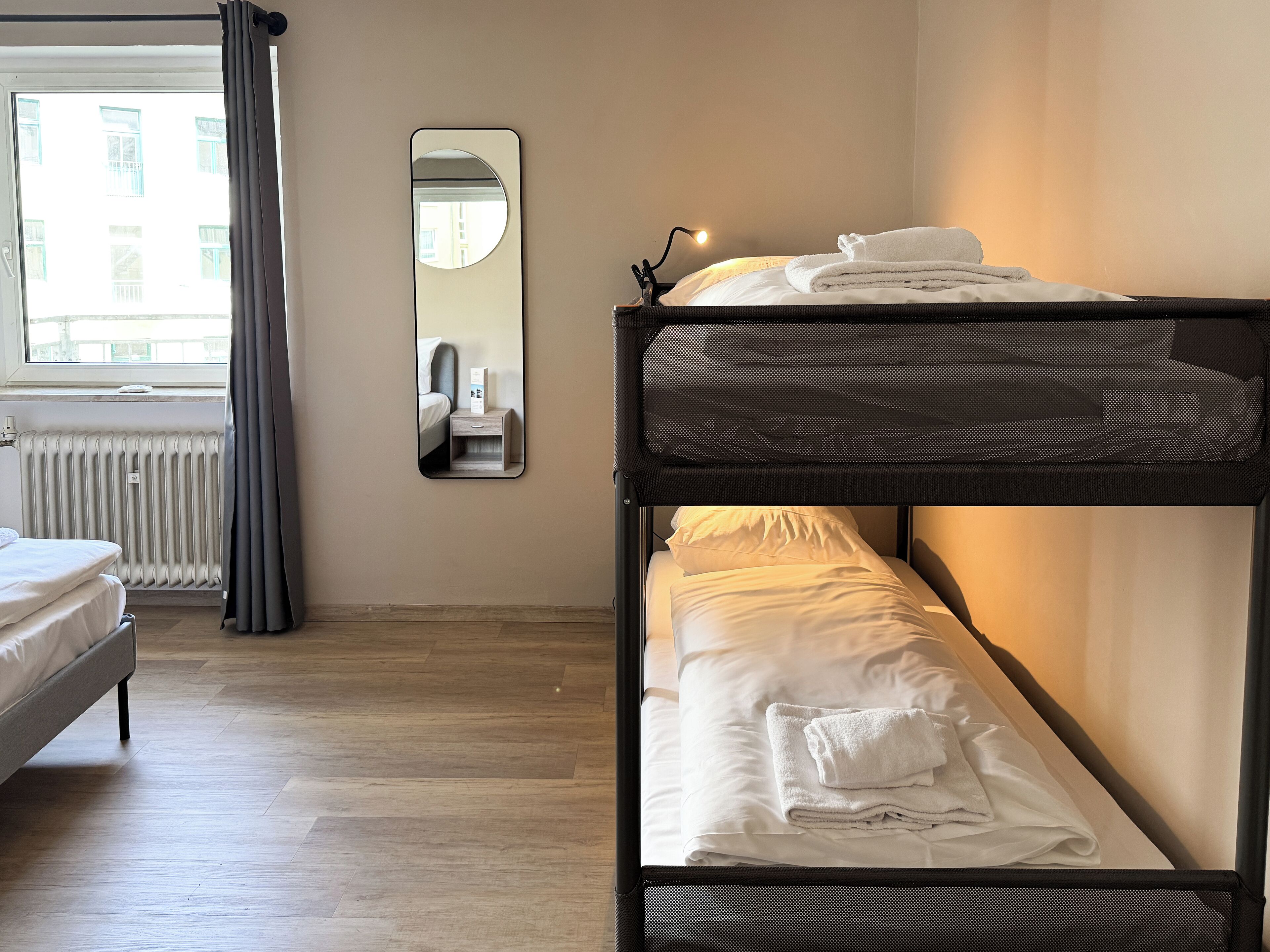 A room with two bunk beds, a window with curtains, a radiator, a mirror, and a bedside table with a lamp.
