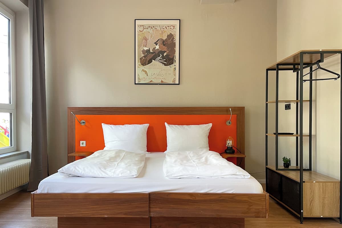 Ein Hotelzimmer mit einem hölzernen Bett, weißer Bettwäsche, einem orangefarbenen Kopfteil, einem Poster an der Wand und einem Metallschrank.