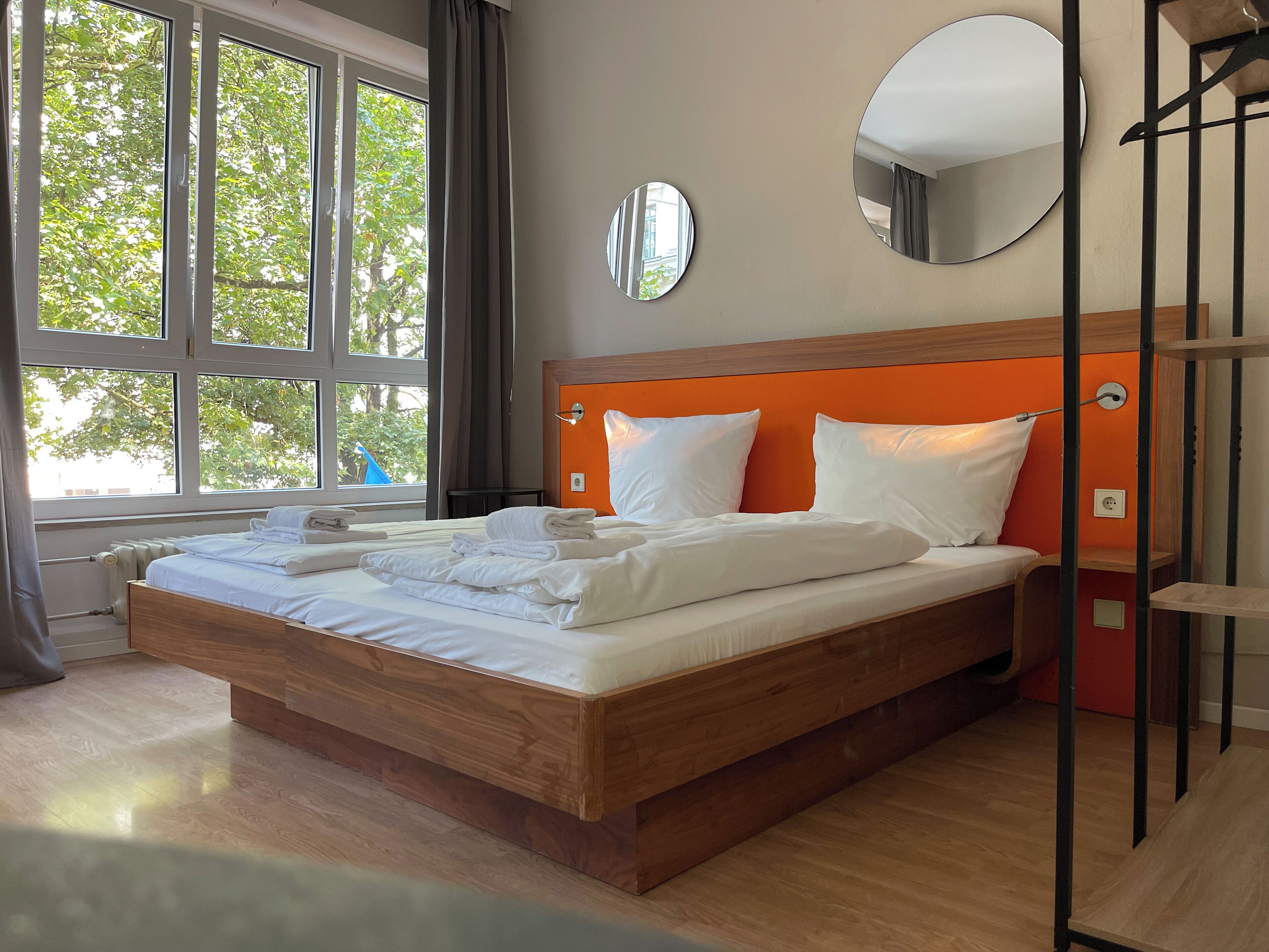A neatly made bed with white linens and towels, a large window with curtains, and a round mirror on the wall.