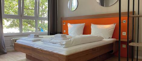 Een opgemaakt bed met witte lakens en handdoeken, een groot raam met gordijnen, en een ronde spiegel aan de muur.