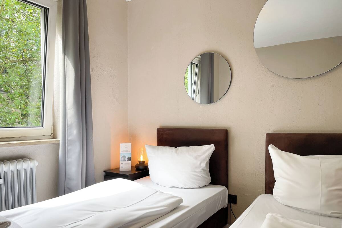 Ein Hotelzimmer mit zwei Betten, einem runden Spiegel, einem Nachttisch mit Lampe und einem Fenster mit Vorhängen.