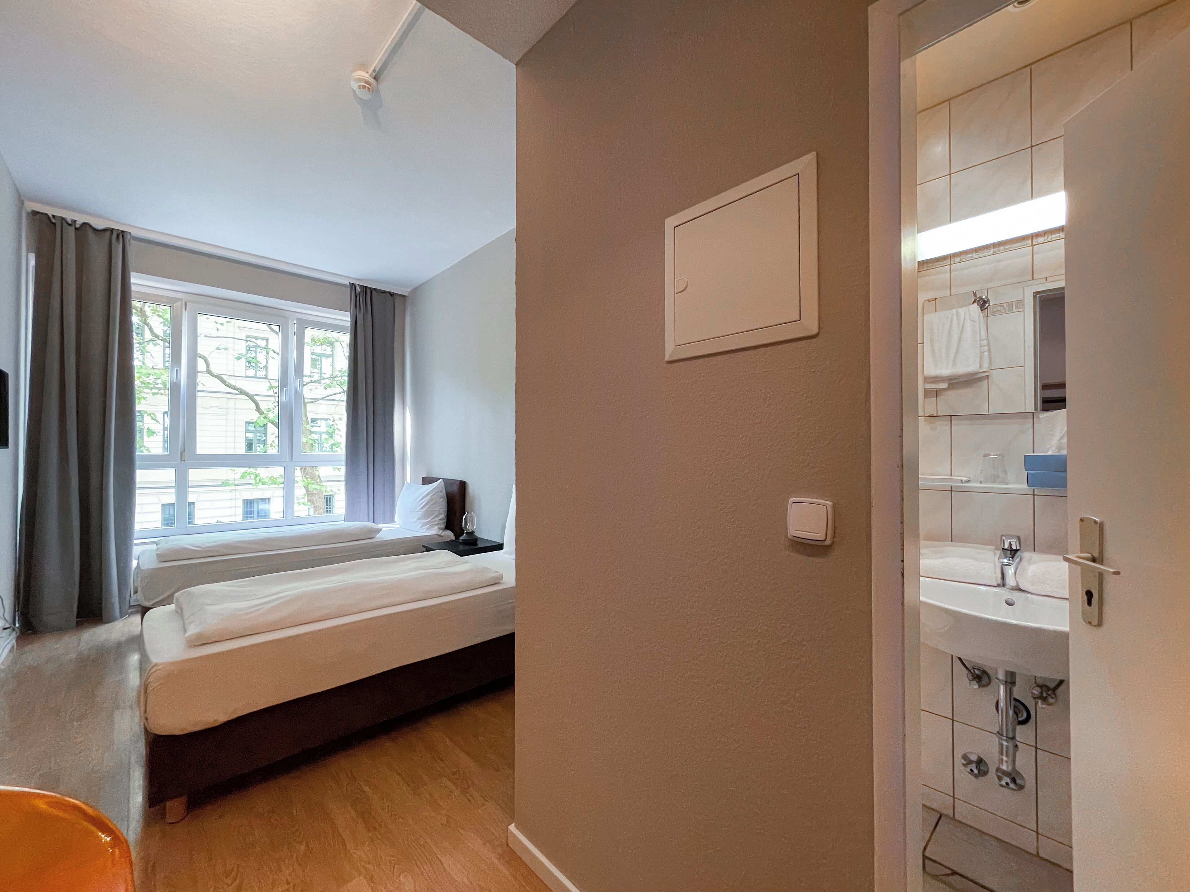 A hotel room with a bed, a window with curtains, a wall-mounted light switch, and a bathroom with a sink and mirror.