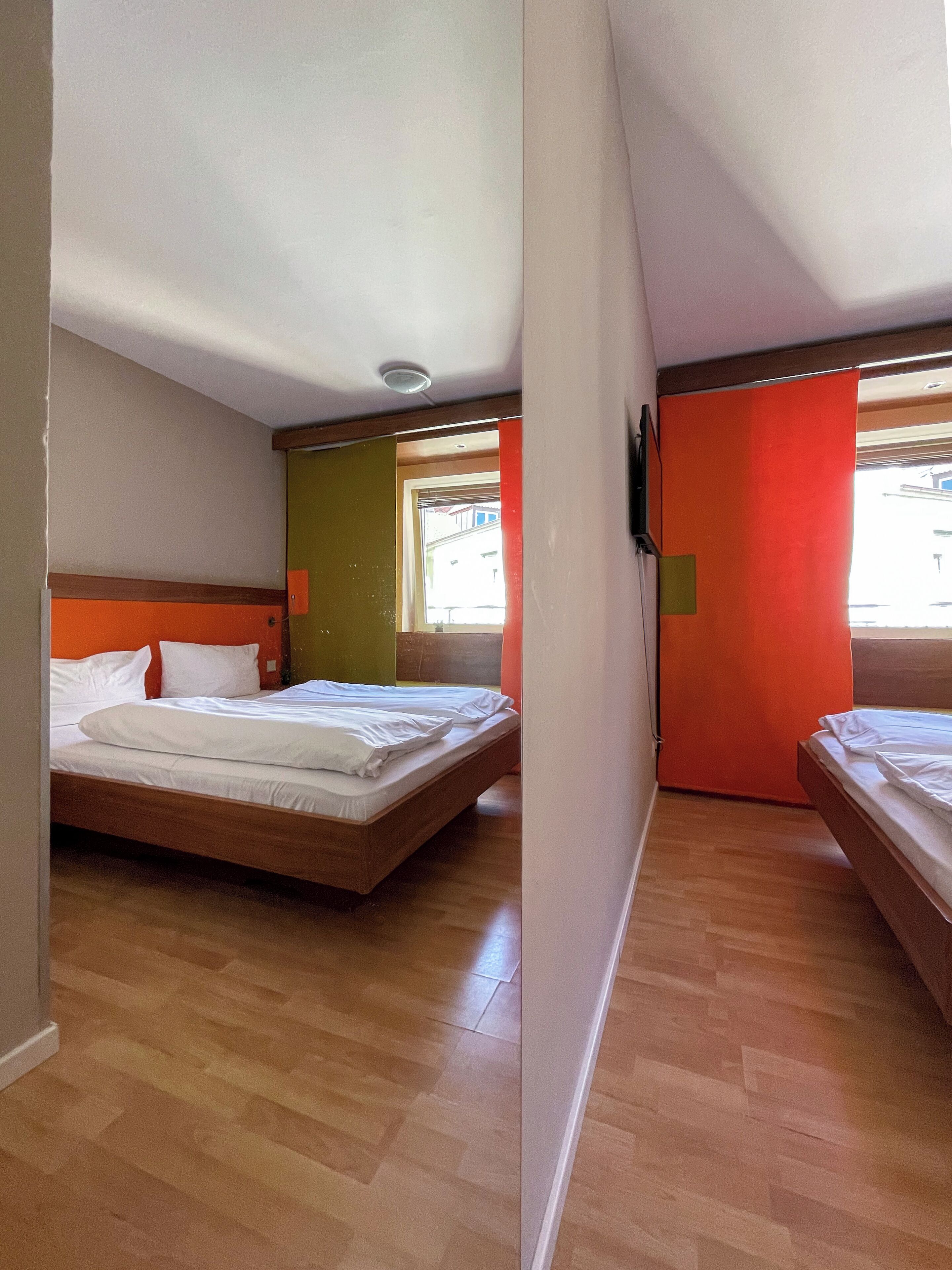 Une chambre d’hôtel avec deux lits, un plancher en bois, une fenêtre et une porte avec un rideau rouge.