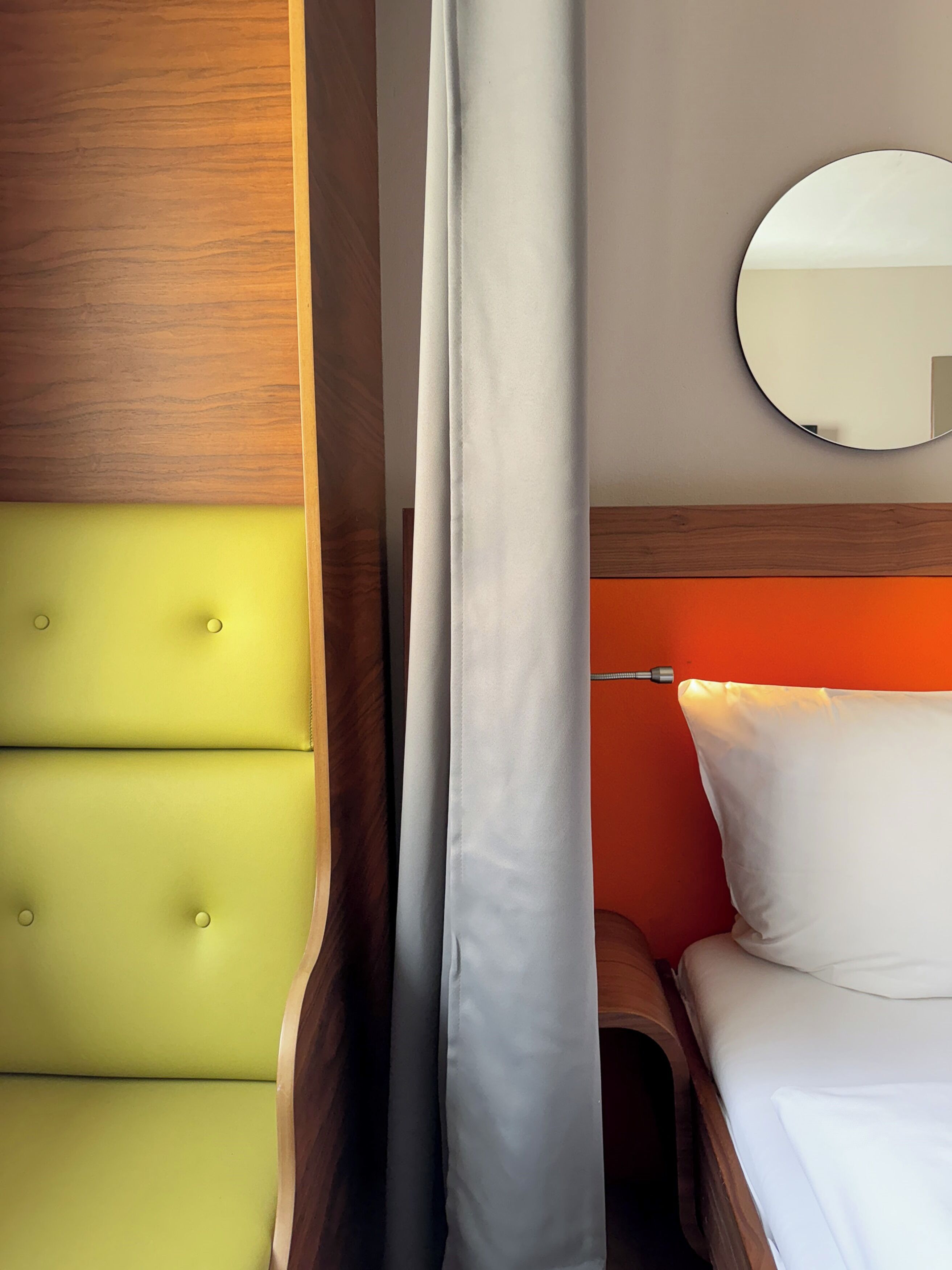 A room with a green upholstered headboard, a wooden headboard, a white curtain, a round mirror, and a white pillow.