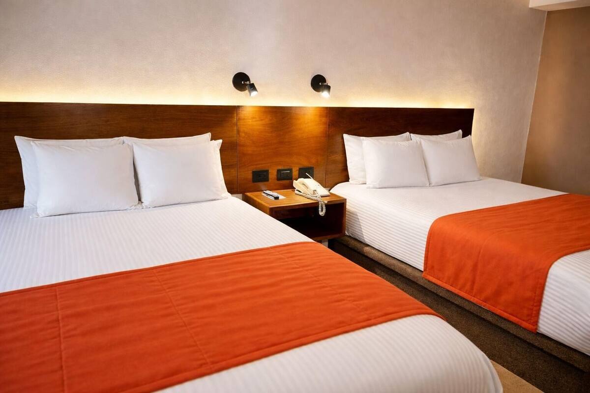 A hotel room with two beds, each with white bedding and orange throw blankets. There are bedside tables with lamps and a phone.