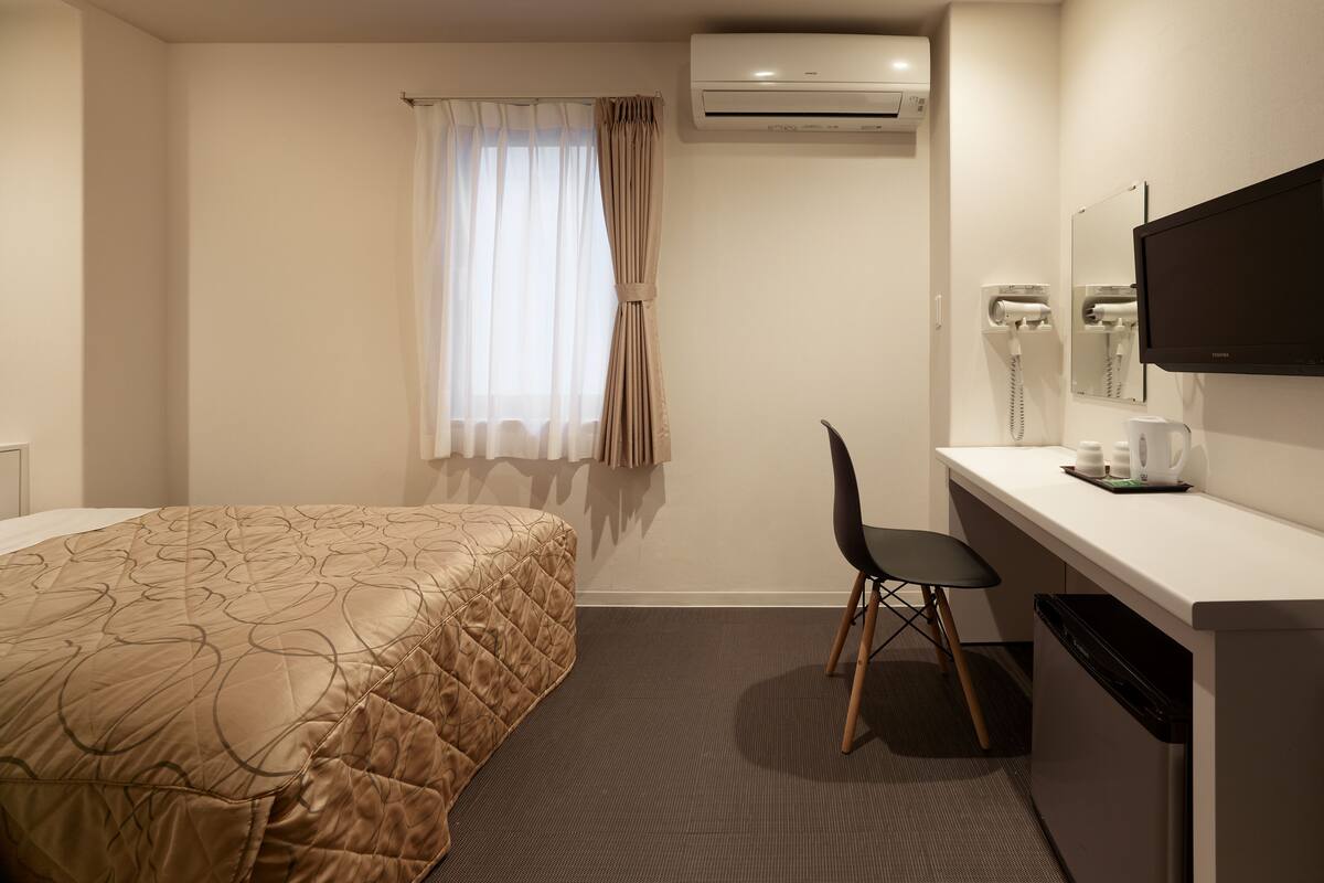 A hotel room with a bed, a desk, a chair, an air conditioning unit, a television, and a hairdryer.