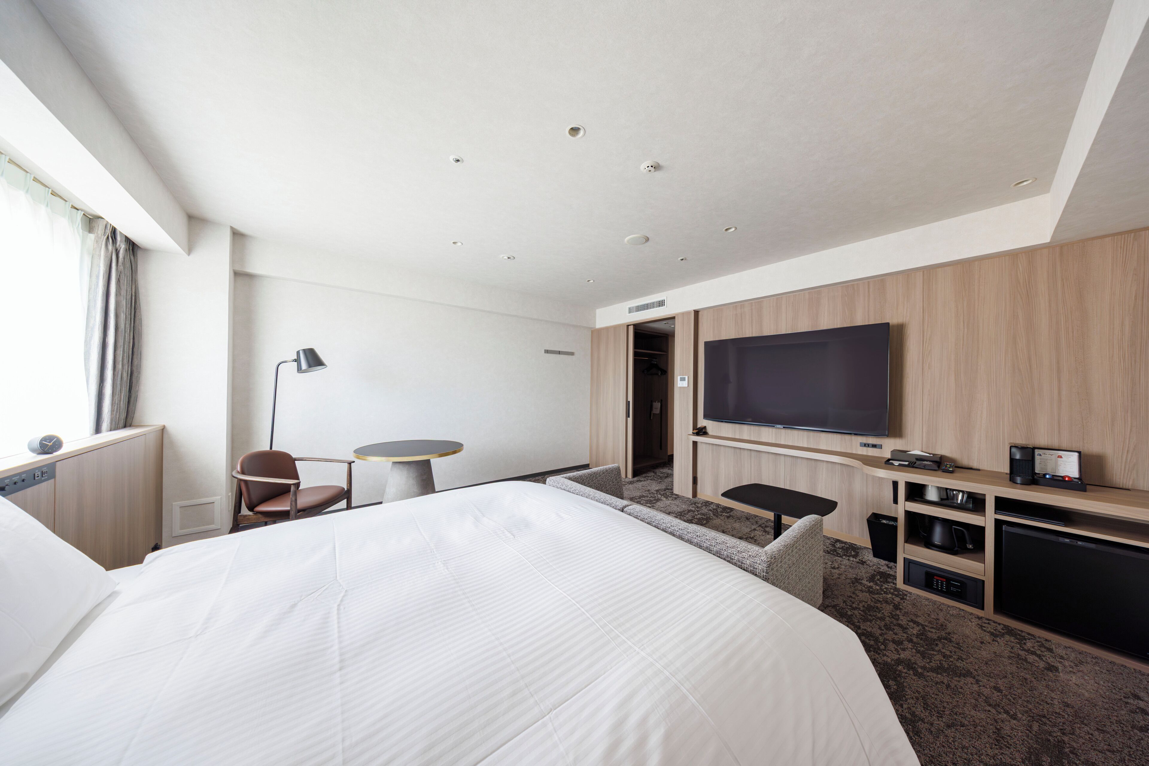 A hotel room with a large bed, a flat-screen TV mounted on the wall, a desk with a chair, and a small seating area with a sofa.