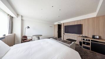 A hotel room with a large bed, a flat-screen TV mounted on the wall, a desk with a chair, and a small seating area with a sofa.