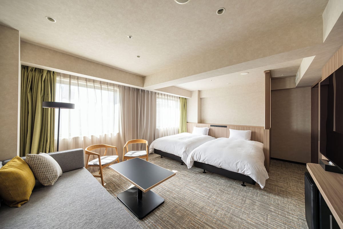 Ein Hotelzimmer mit zwei Betten, einem Sofa, einem Tisch, einem Fernseher und einem fenster mit Vorhängen.