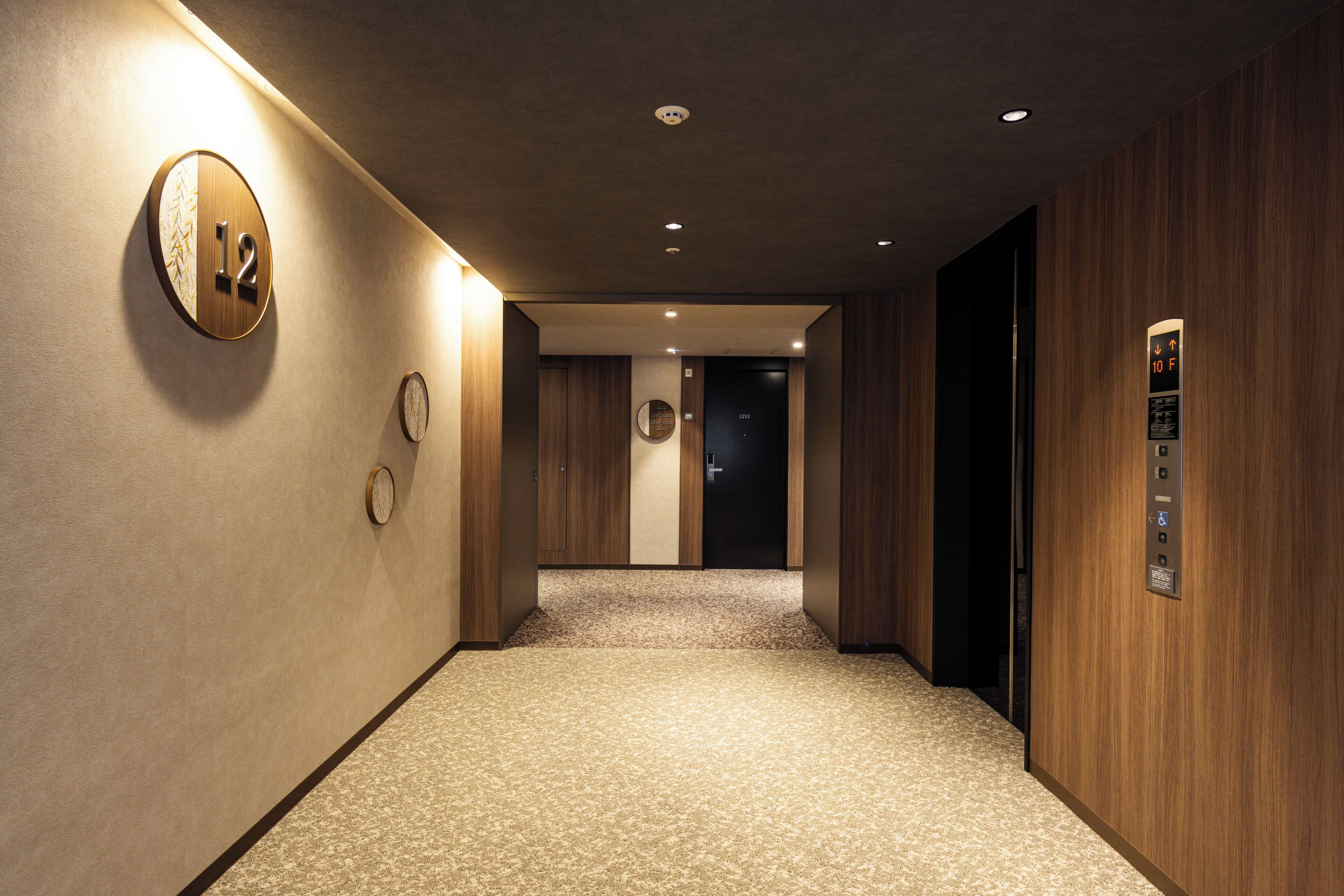 A hotel hallway with a wooden wall, a carpeted floor, a round wall clock, and an elevator.