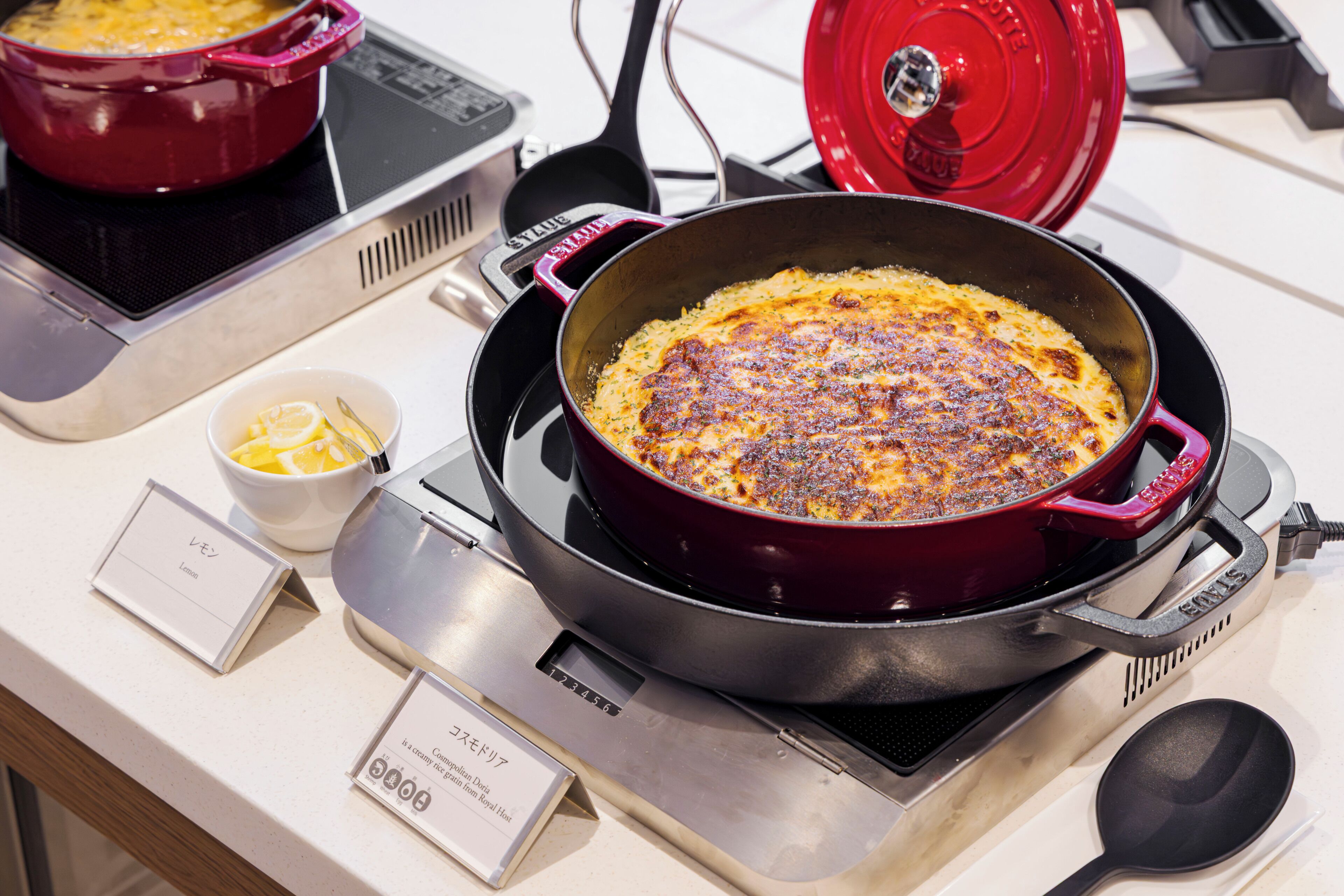 A red pot on a stove with a lid, a bowl of lemon slices, and a spoon on a table.