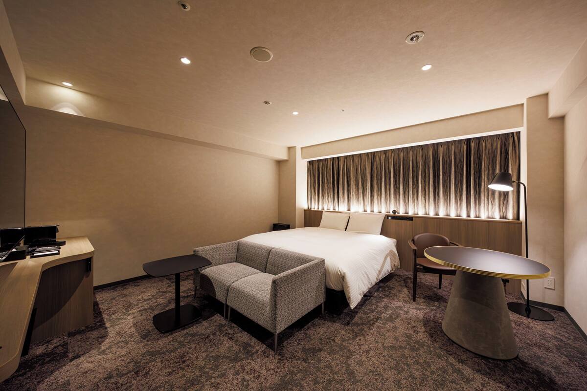 Ein Hotelzimmer mit einem Bett, einem Schreibtisch, einem Stuhl, einem kleinen Tisch und einem Fenster mit Vorhängen.