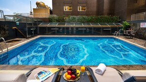 Una piscina con diseño de mosaicos, rodeada de sillas de descanso y una mesa con fruta y una toalla.