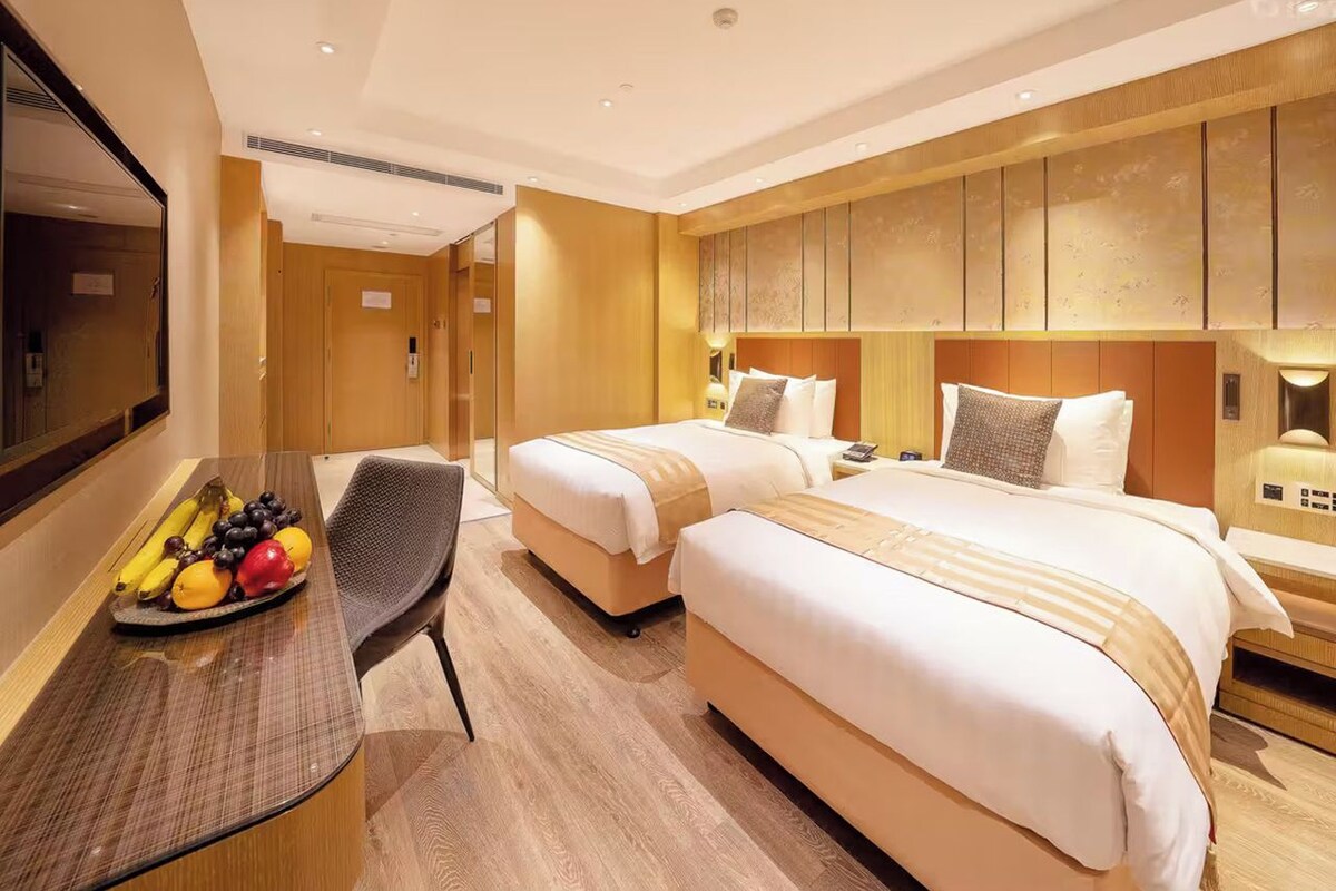 A hotel room with two beds, a desk with a fruit bowl, and a mirror.