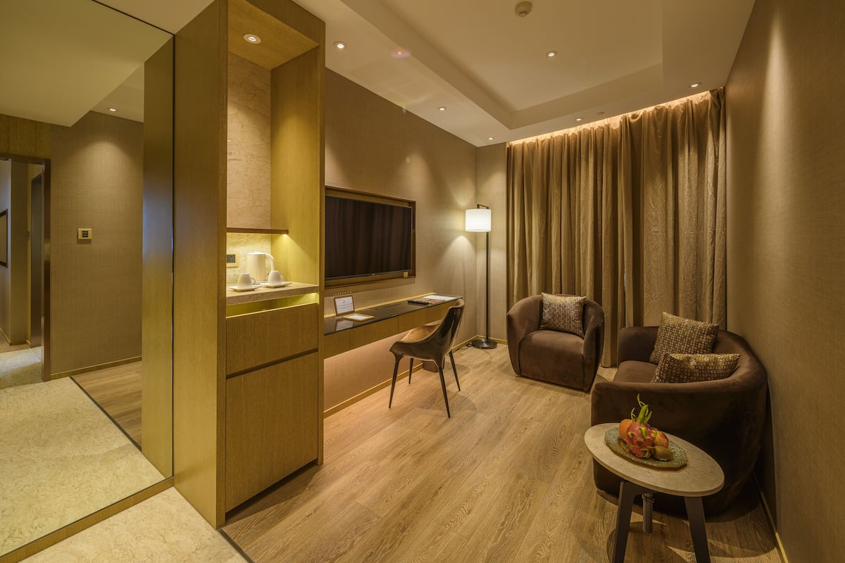 A modern hotel room with a wooden floor, a desk with a laptop, a sofa, a small table with a fruit bowl, and a TV mounted on the wall.