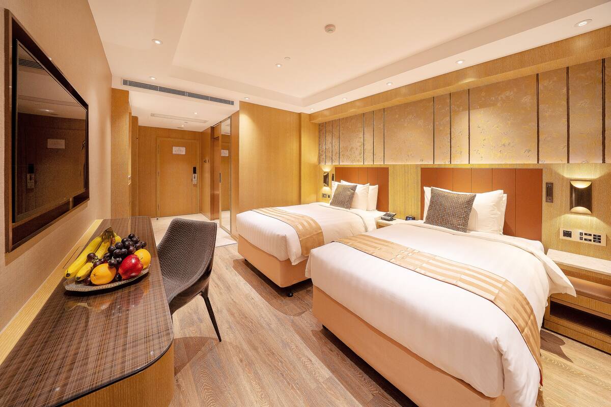 A hotel room with two beds, a desk with a fruit tray, and a large mirror.