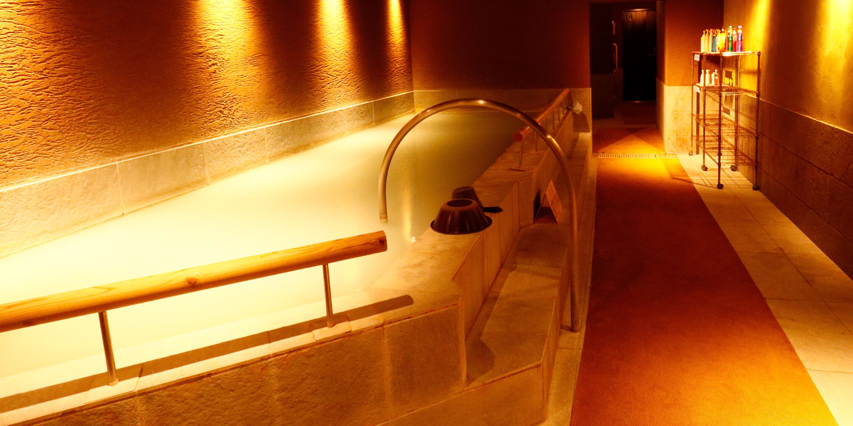 A hallway with a glass-enclosed bathtub, a wooden handrail, and a metal rack with bottles.