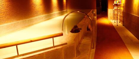 A hallway with a glass-enclosed bathtub, a wooden handrail, and a metal rack with bottles.